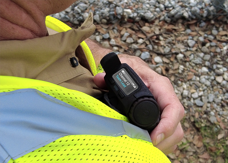 Worker in HiVis vest holding TAIT AXIOM Wearable device looking down at the heads-up display screen. 